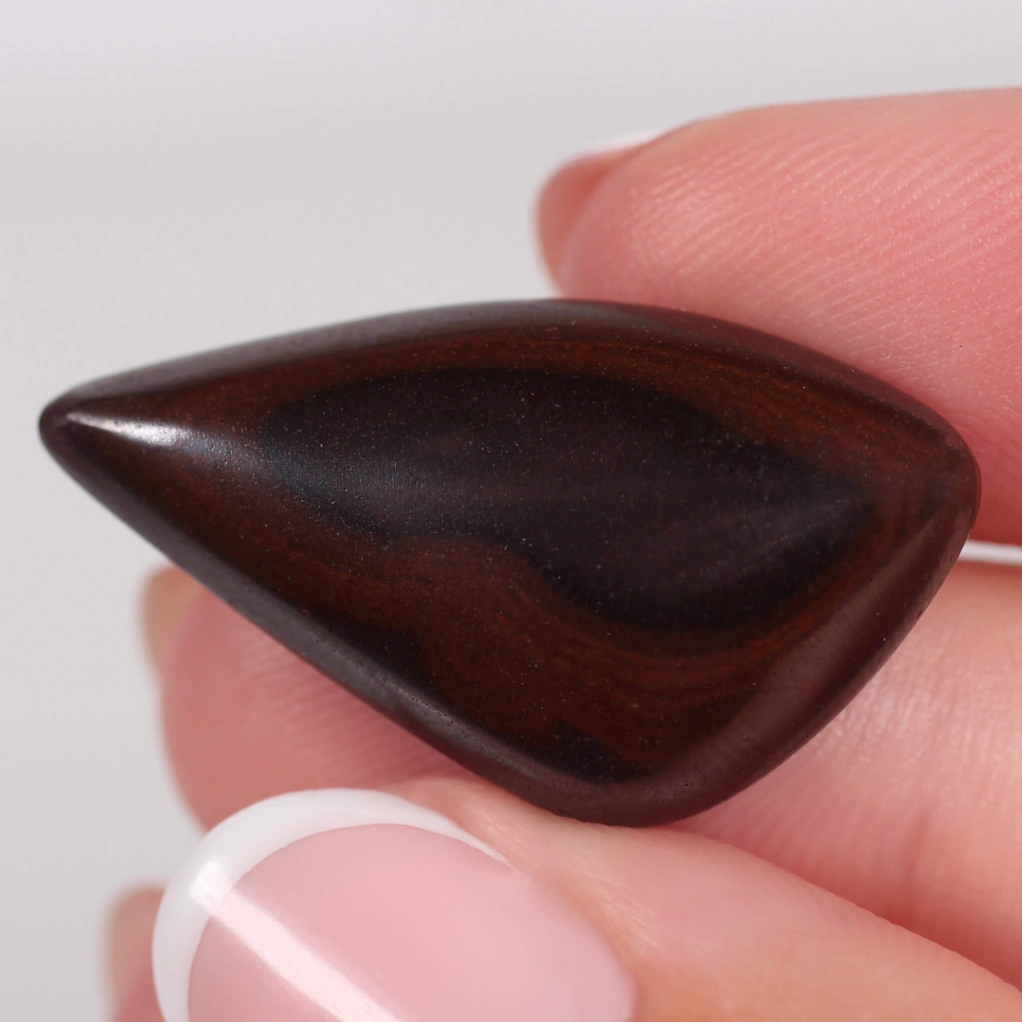 Teardrop-shaped dark brown stone held between fingers against a white background